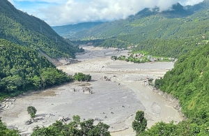 Überschwemmtes Flusstal in Nepal mit Schlammmassen und beschädigten Gebäuden zwischen bewaldeten Berghängen.