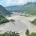 Überschwemmtes Flusstal in Nepal mit Schlammmassen und beschädigten Gebäuden zwischen bewaldeten Berghängen.