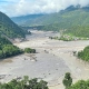 Überschwemmtes Flusstal in Nepal mit Schlammmassen und beschädigten Gebäuden zwischen bewaldeten Berghängen.