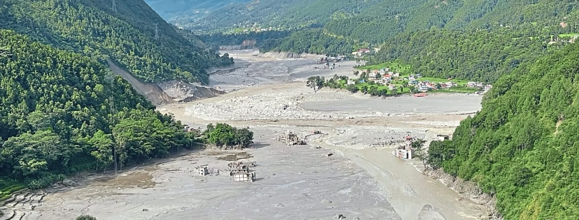 Überschwemmtes Flusstal in Nepal mit Schlammmassen und beschädigten Gebäuden zwischen bewaldeten Berghängen.